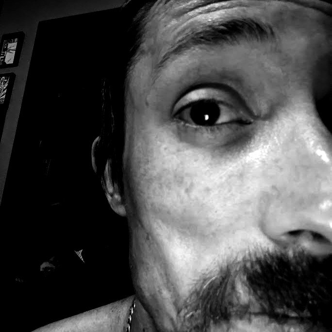 A close-up selfie of Derjyn, cut off by the frame: one eye on the camera, a short beard, a dark room behind, printed in ordered dither.